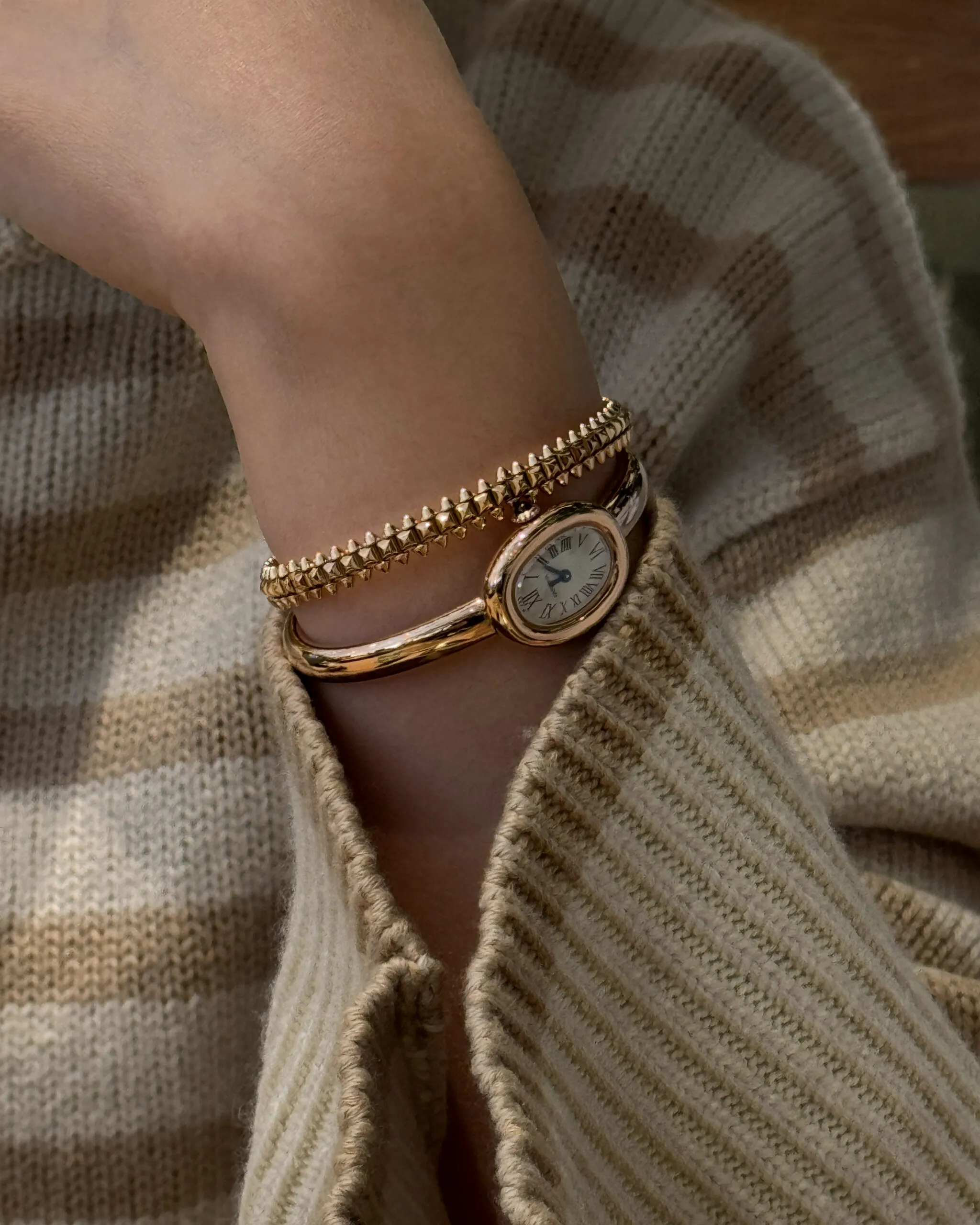 A gold oval Cartier watch worn with a studded gold bracelet over a cream knit sleeve