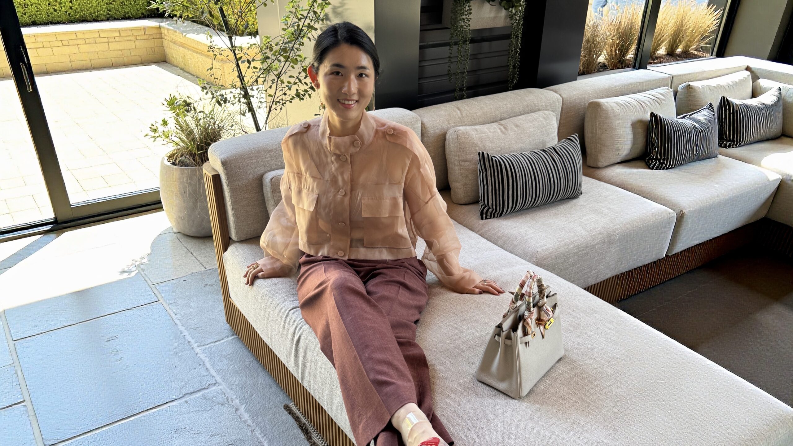 Lily sitting on an outdoor sofa wearing a sheer blush jacket and burgundy trousers, styled with a Hermès Birkin bag
